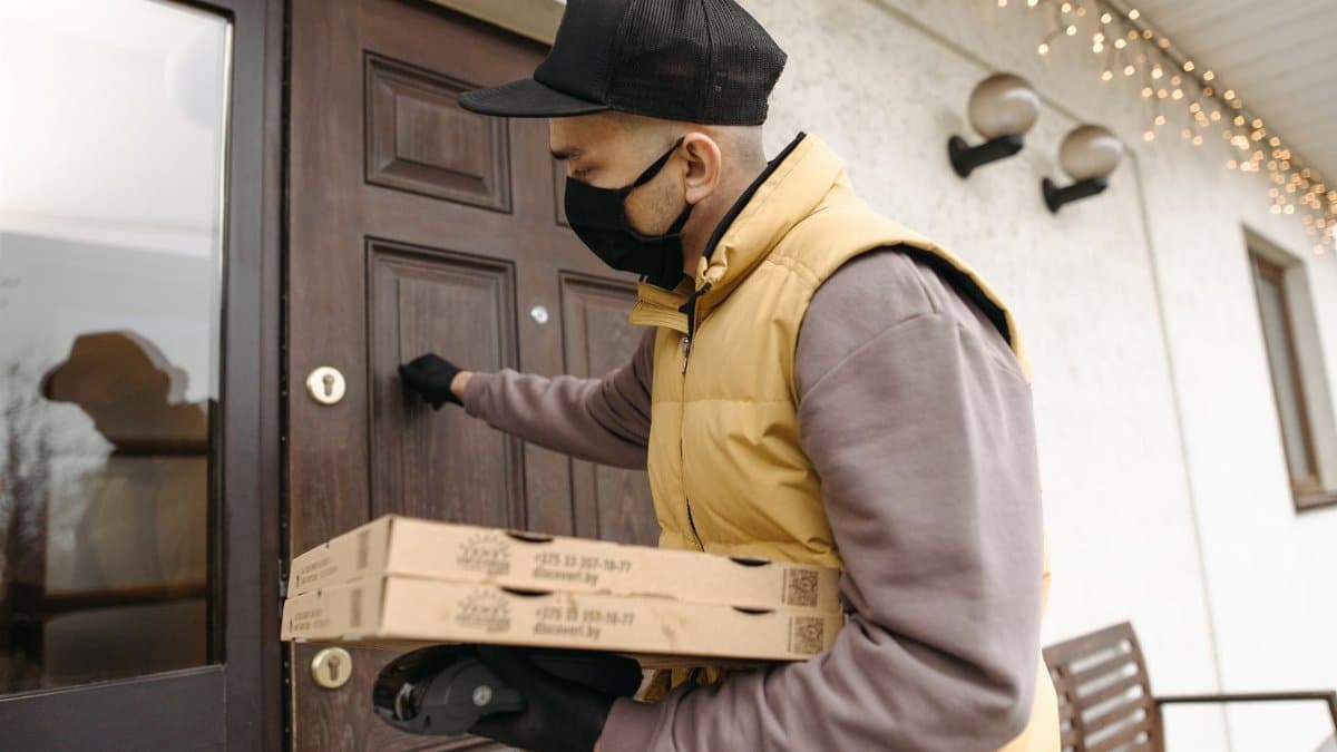 A deliveryman wearing a mask and gloves knocks on a door while holding pizza boxes, emphasizing safety during delivery.