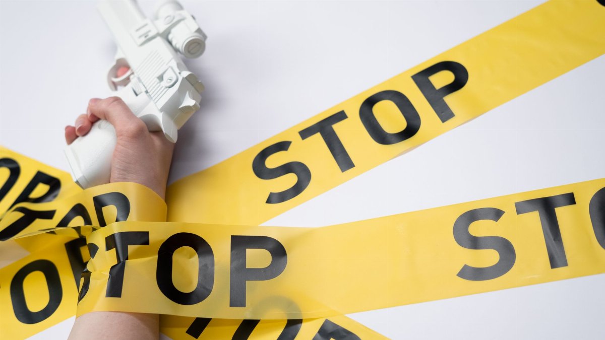 A white toy gun held by a hand wrapped in yellow stop tape on a white background, conveying a message of caution.