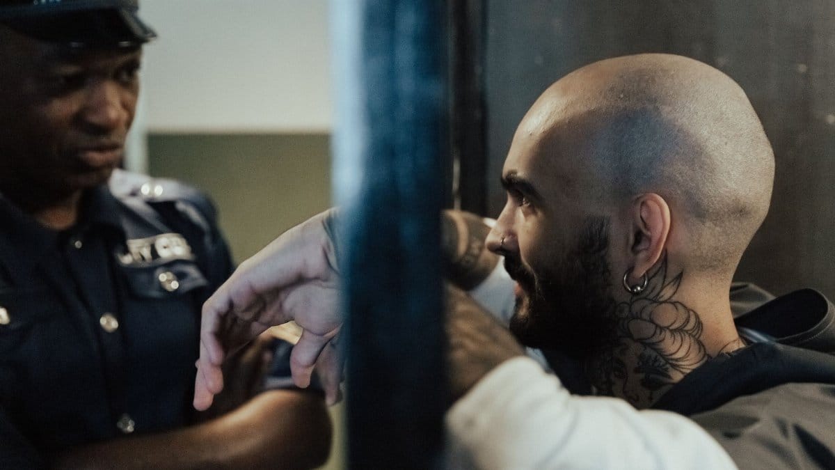 Close-up of a police officer interacting with a tattooed man behind metal bars inside a jail.