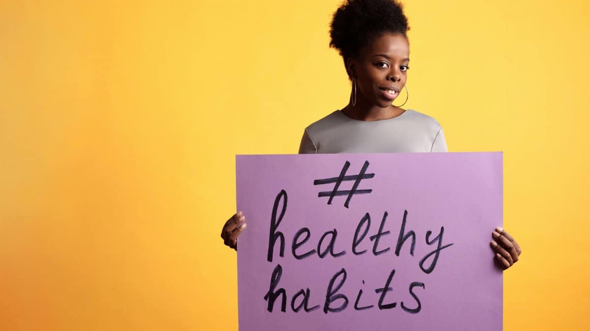 Portrait of a woman holding a healthy habits sign, set against a vibrant yellow background.