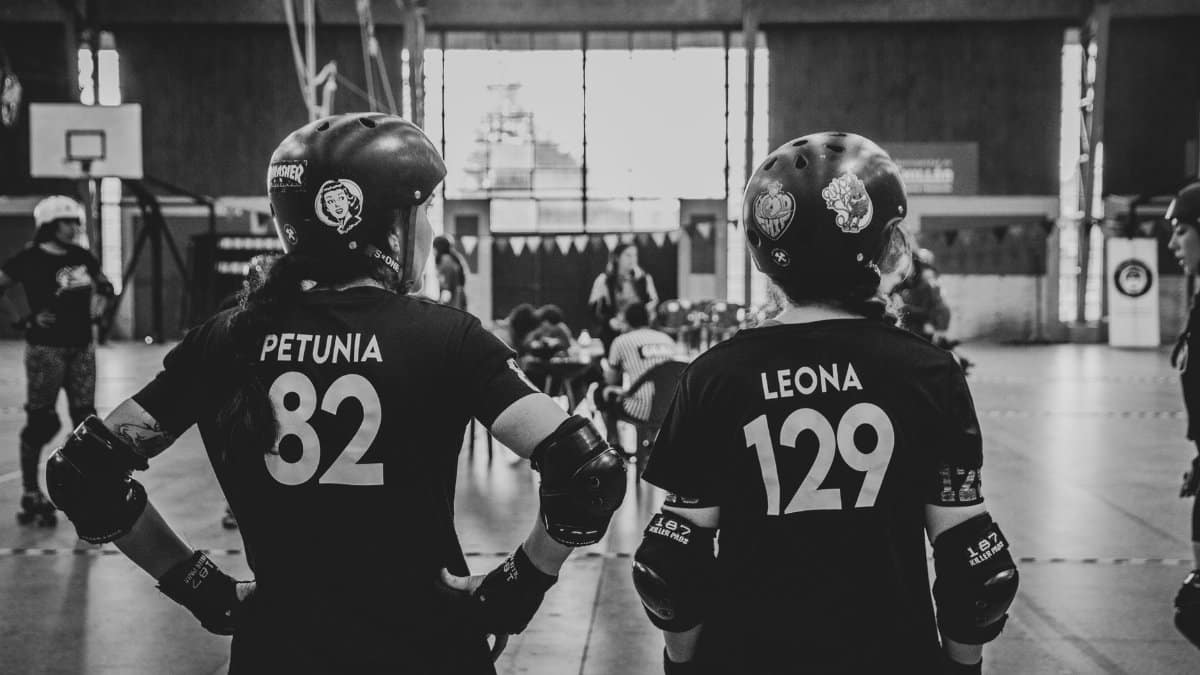 Black and white image of two roller derby women athletes standing in a skatepark wearing helmets and sportswear.