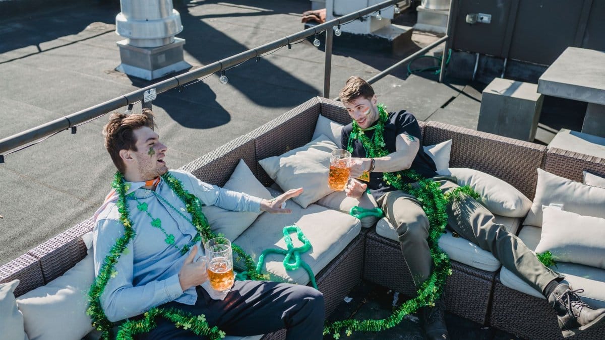 From above of Irish friends sitting on sofa with beer while celebrating holiday on terrace and looking at each other