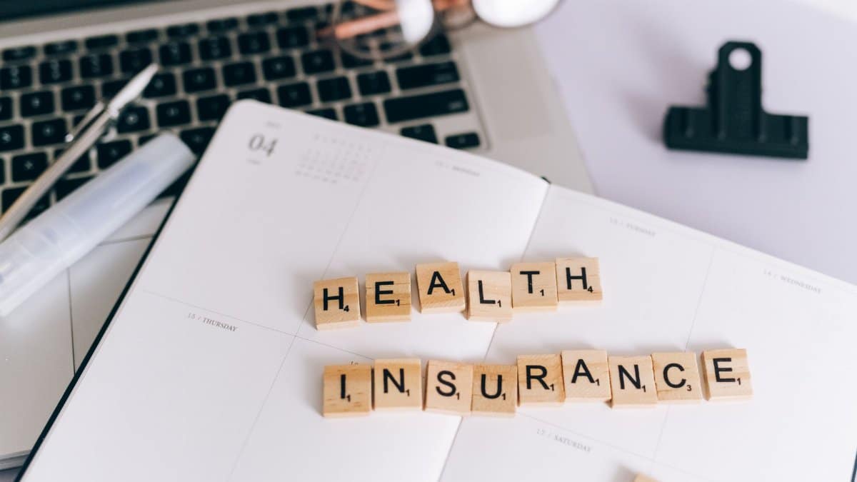 Close-up of Scrabble tiles spelling 'Health Insurance' on a planner with a laptop.