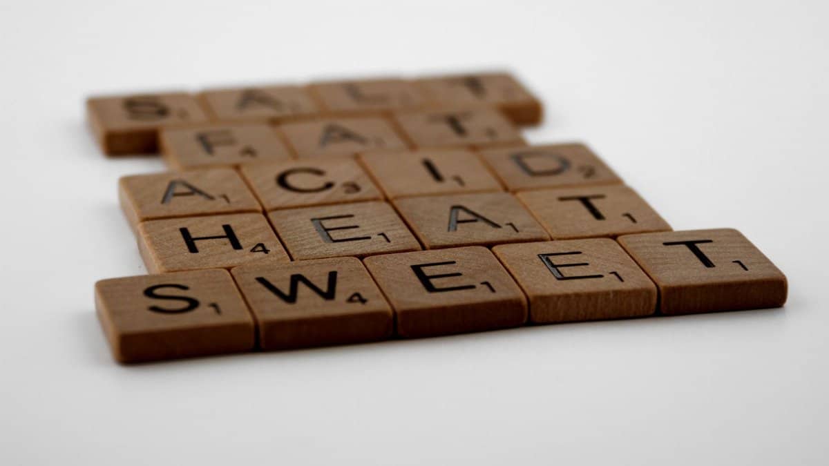 Wooden Scrabble tiles arranged to spell 'Salt, Fat, Acid, Heat, Sweet' on a white background.