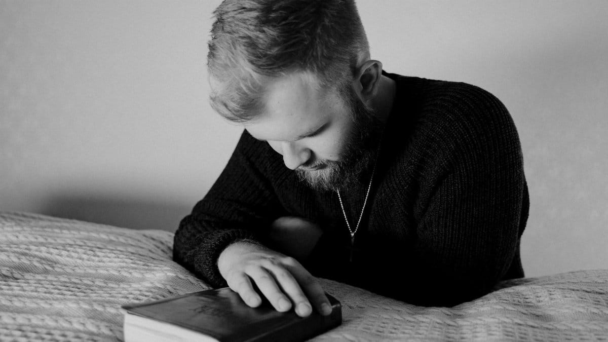 A bearded man in a knitted sweater prays over a Bible, conveying faith and contemplation.