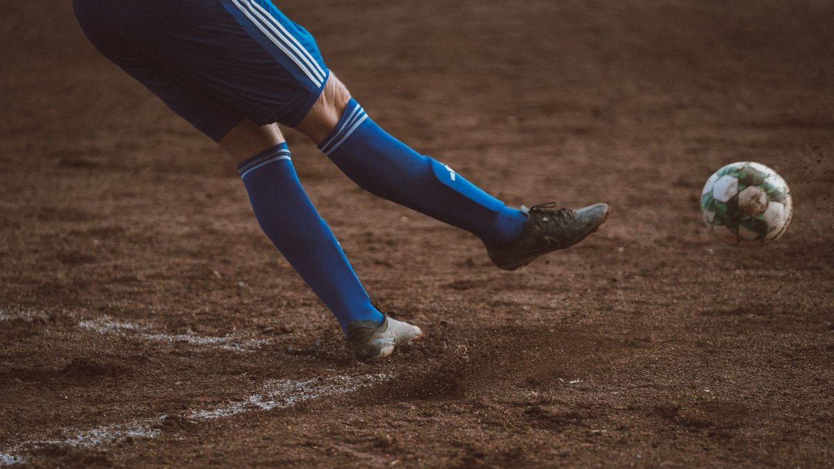Dynamic soccer player kicks ball on outdoor dirt field. Focus on leg action.