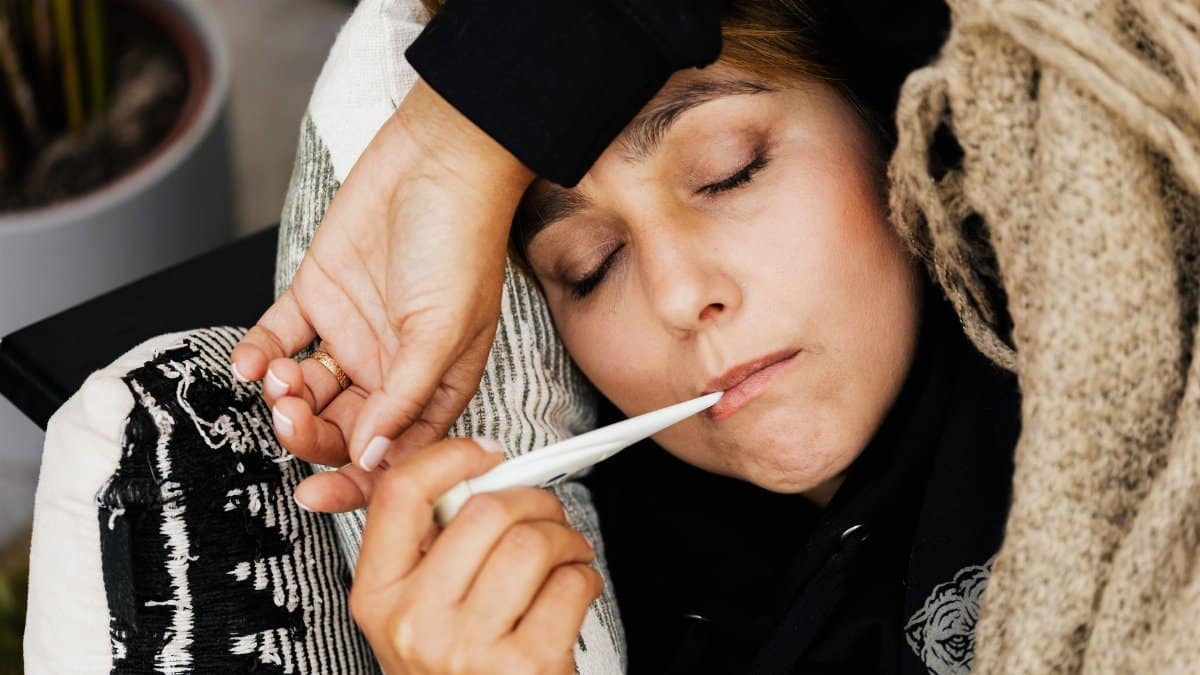 A woman resting on a couch using a thermometer to check her temperature, wrapped in a cozy blanket.
