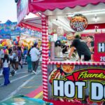Bustling outdoor fair with a popular hot dog stand and diverse crowd.