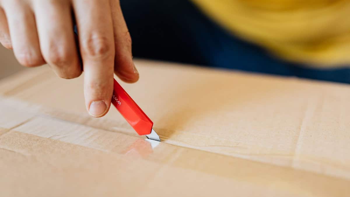 Crop anonymous male unpacking belongings from cardboard box by using sharp utility knife after getting delivery during relocation