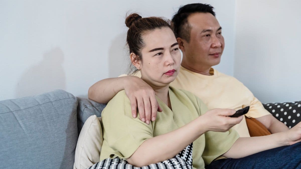 An Asian couple sitting on a couch at home, watching TV together with a remote control in hand.