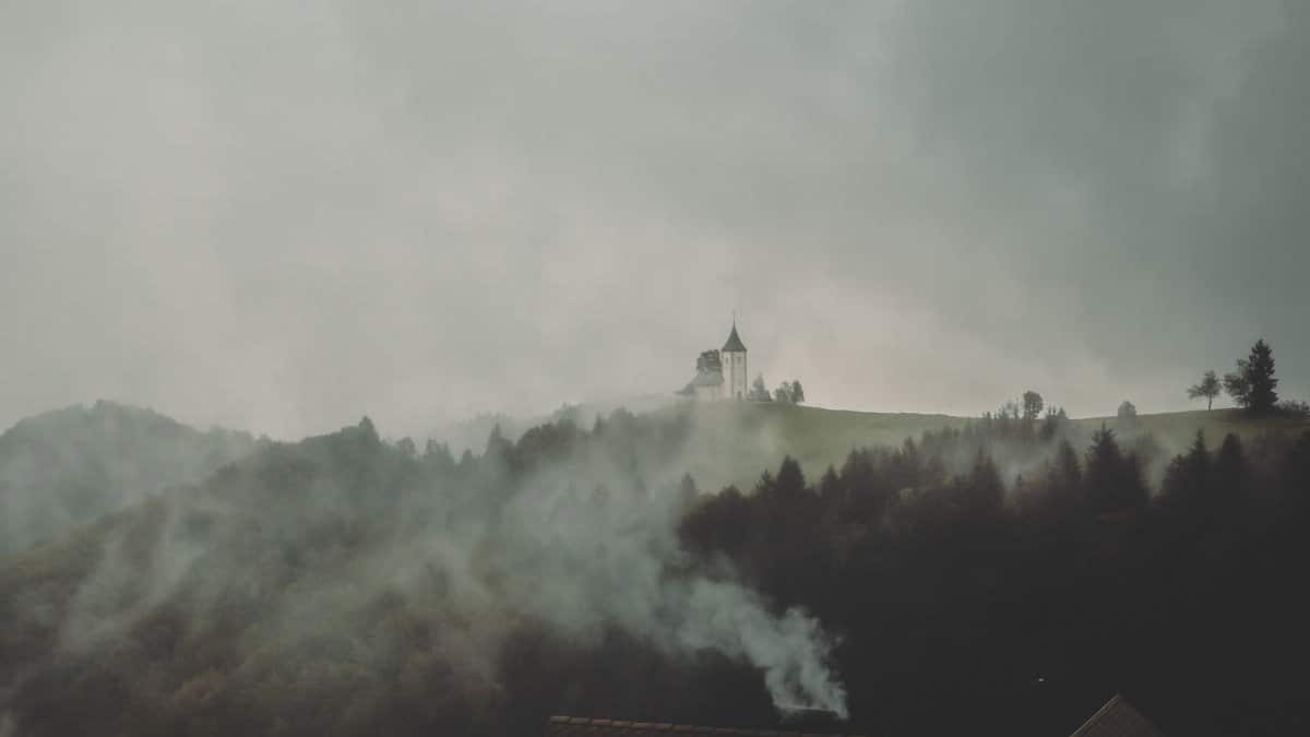 A moody landscape of a distant church on a misty hillside framed by trees and smoke, under a gray sky.