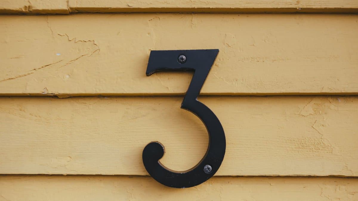 Close-up of a black number three sign mounted on a textured yellow wooden wall.