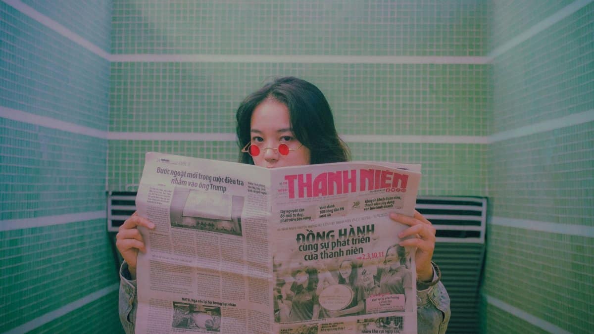 An Asian woman wearing sunglasses, reading a newspaper with a retro feel indoors.