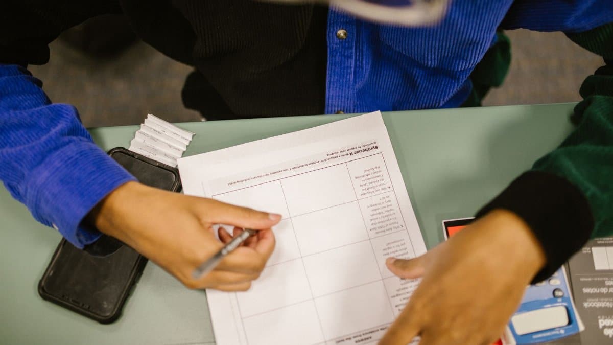 Overhead view of a student writing on a test paper in a classroom setting with a smartphone nearby.