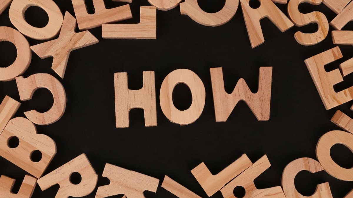 Top view of scattered wooden letters with 'HOW' centered on a dark surface, promoting inquiry.
