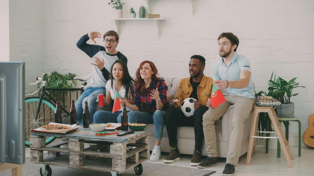 Group of friends cheering while watching a soccer game on TV indoors, holding snacks and flags. Excited atmosphere.