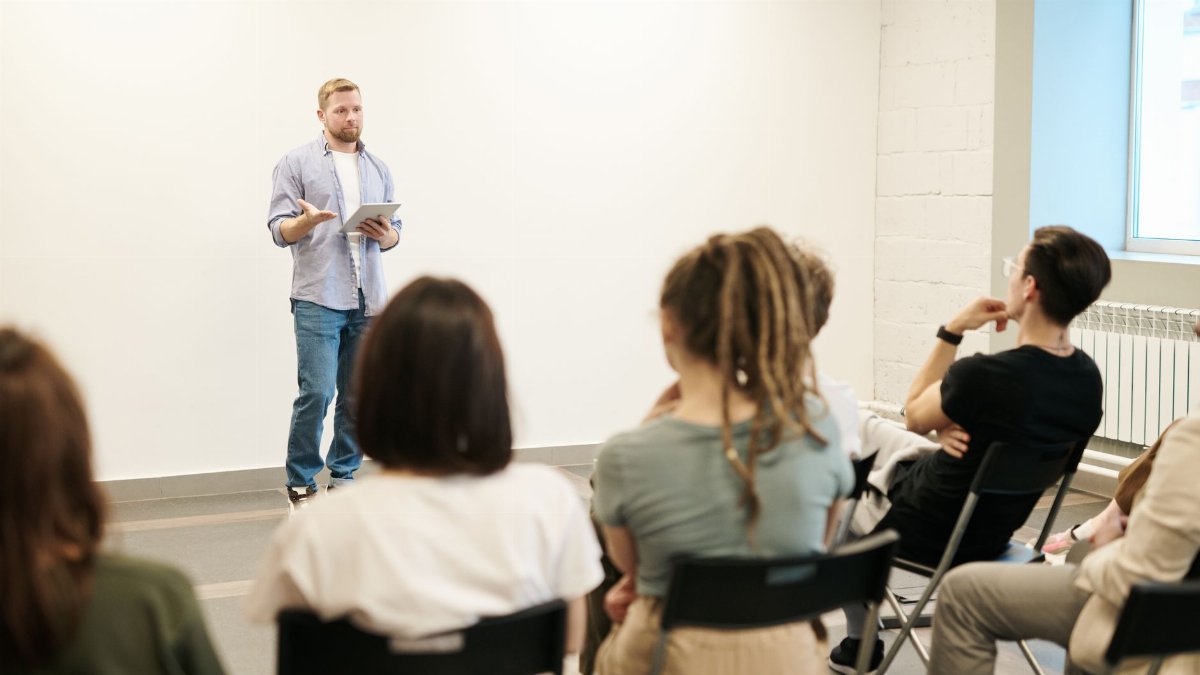 Man presenting with tablet to a small seated audience in a modern indoor setting.