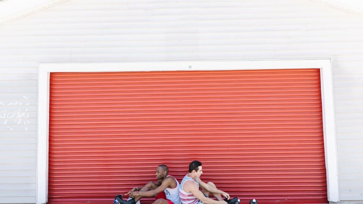 Two adults enjoying outdoors while sitting with roller skates by a red shutter.