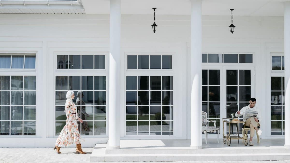A man reads on a porch while a woman in a floral dress walks by. Modern exterior decor.