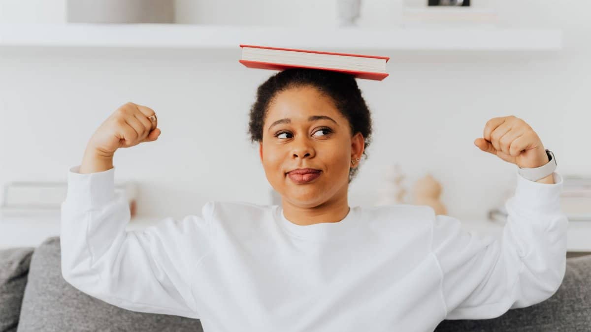 A confident woman balances a book on her head and flexes indoors. Casual and relaxed atmosphere.