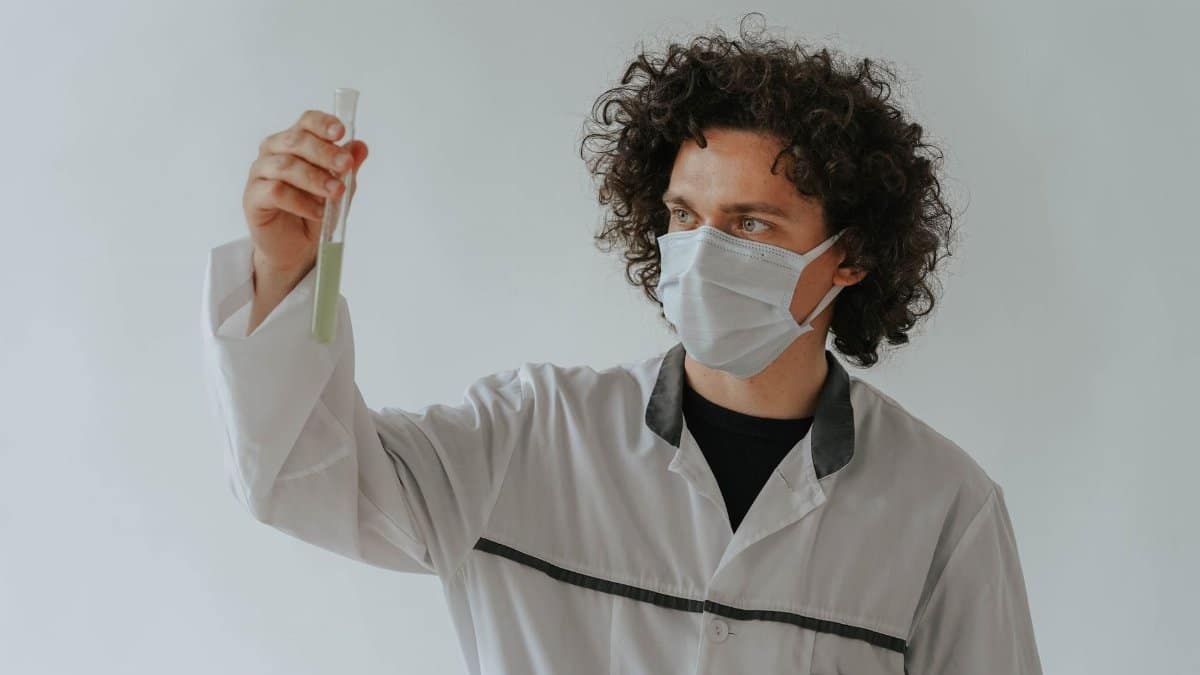 A scientist in a lab coat examining a test tube while wearing a face mask.