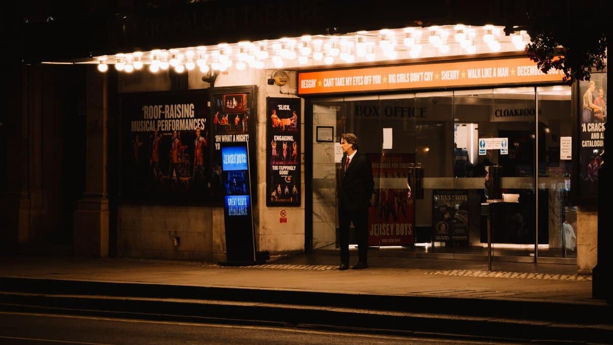 A man waits outside a brightly lit theater entrance on a city street at night, capturing urban elegance.