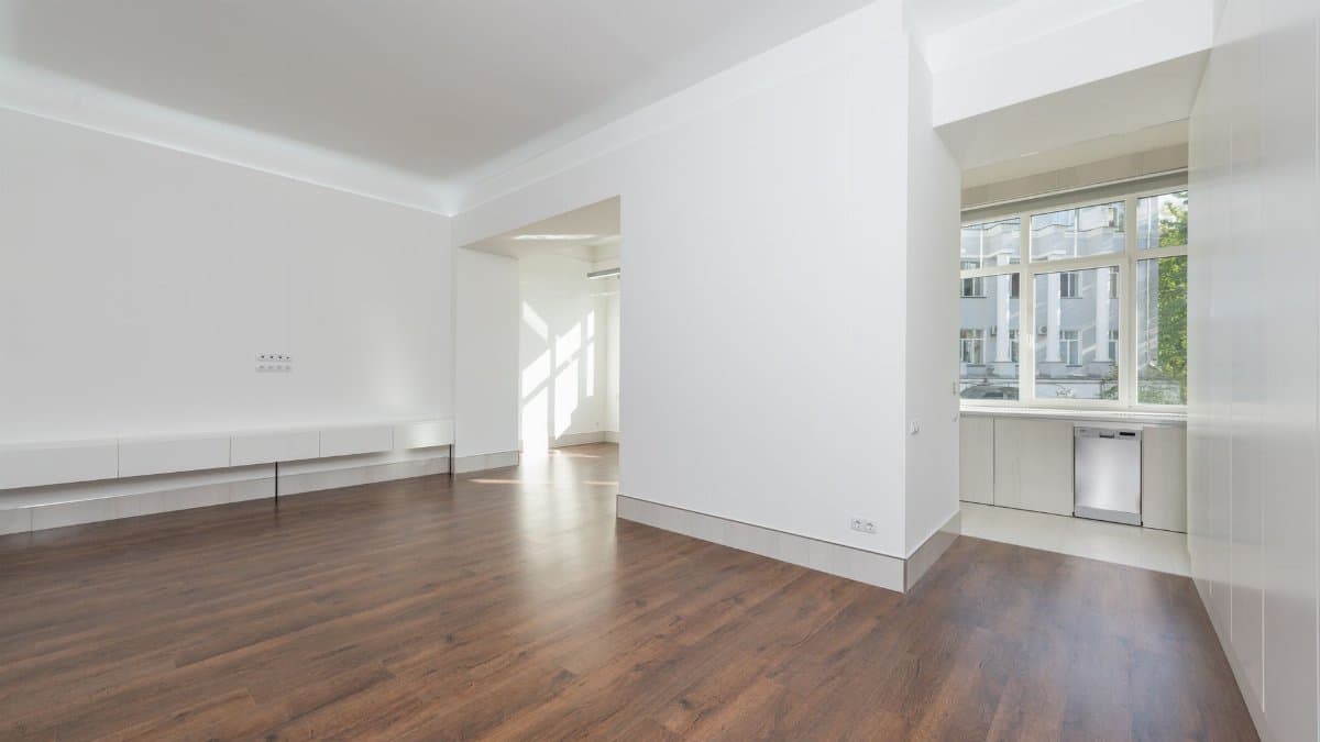 A contemporary empty apartment with white walls and wooden flooring, featuring large windows.