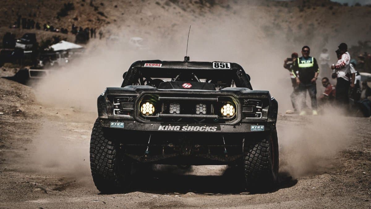High-speed off-road racing action in the Mexicali desert with dust clouds and spectators.