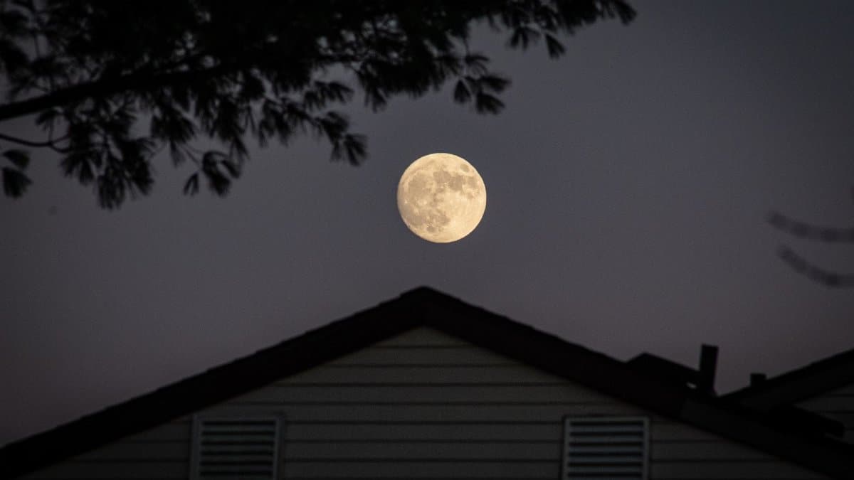 A full moon illuminating the night sky above a silhouetted house with trees.