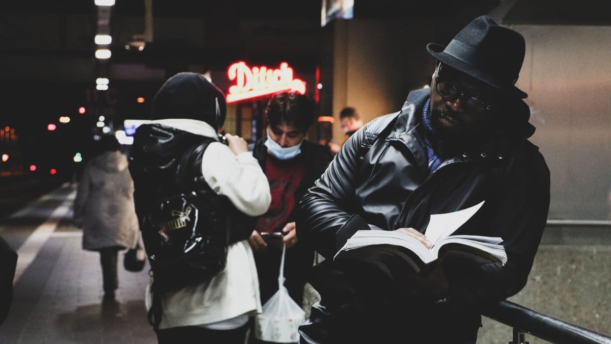 A man in a black leather jacket reads a book on a busy urban street at night.