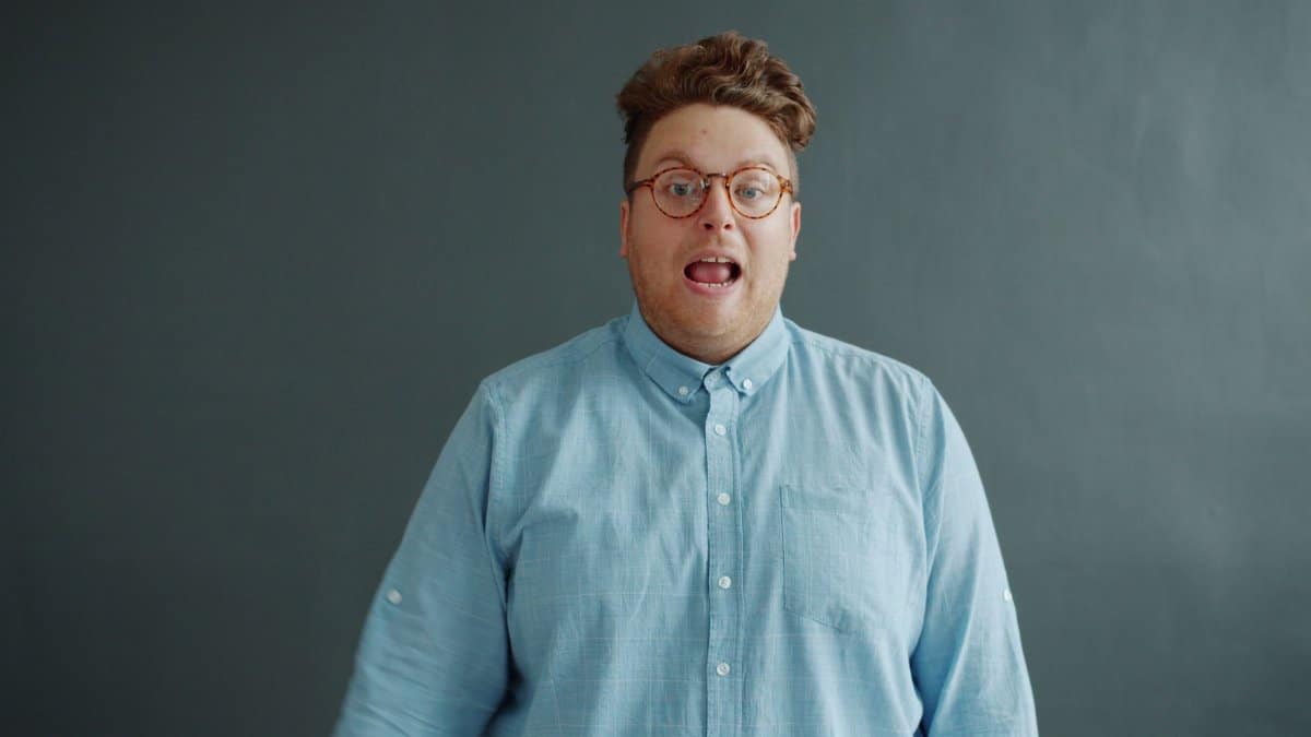 Portrait of a surprised man in glasses and blue shirt against a simple backdrop.