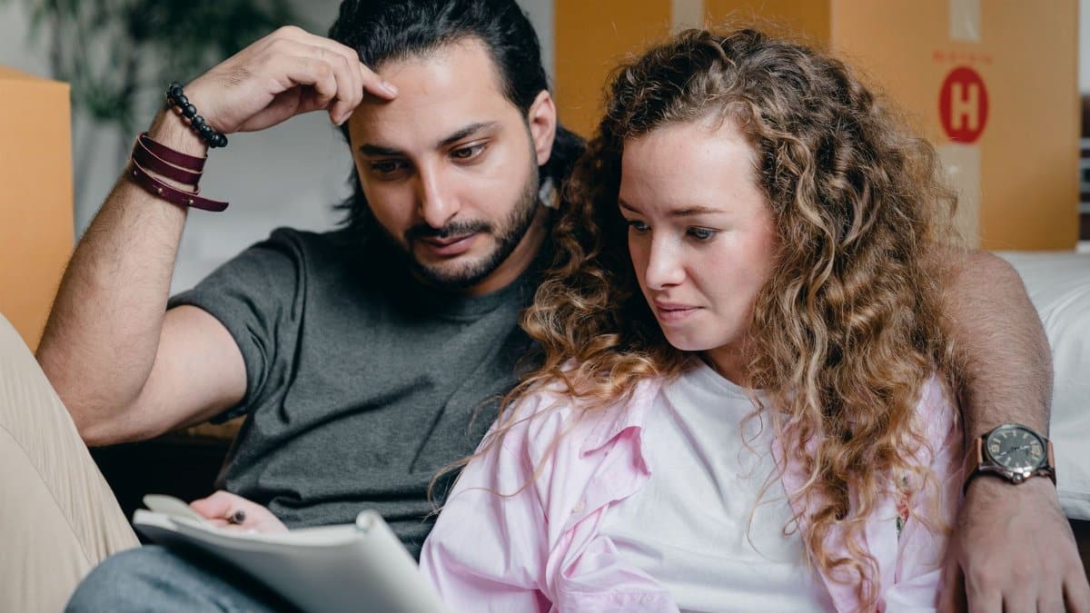 Focused male and female in casual wear sitting near boxes and looking through notebook thoughtfully while moving house