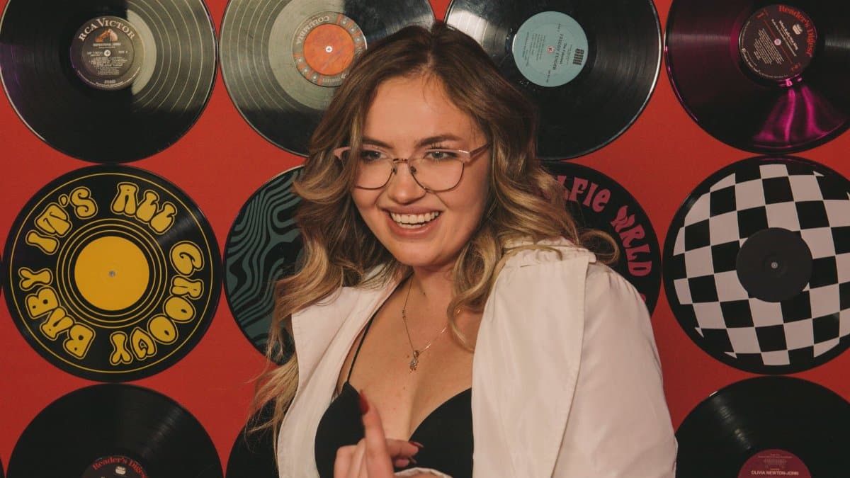 Smiling woman with long hair and eyeglasses posing in front of vinyl record wall.