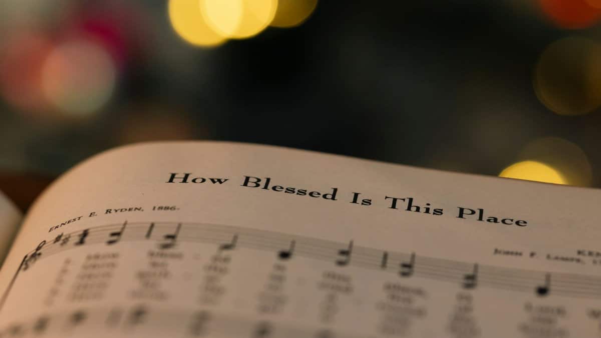 Close-up of a hymn book page with 'How Blessed Is This Place' amidst warm bokeh lights.
