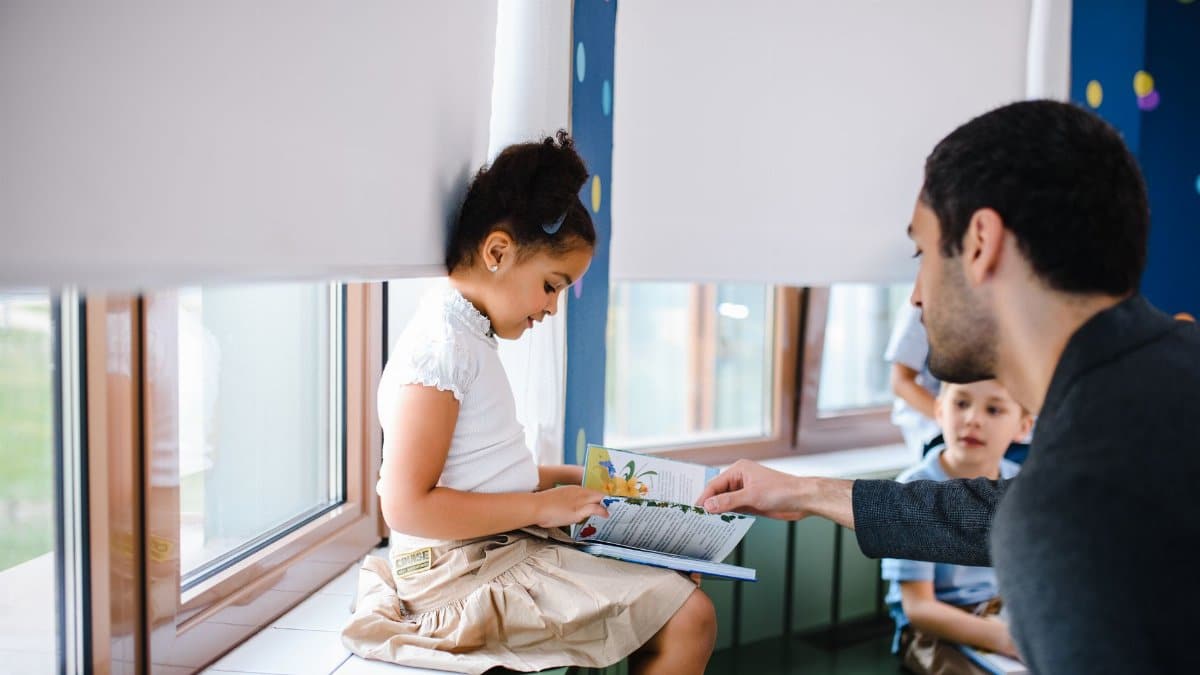A teacher helps a young girl read a book with fellow students in a classroom setting.