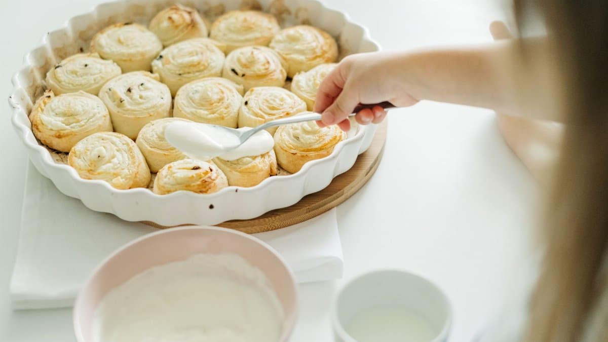A child spreads creamy icing over freshly baked cinnamon rolls, highlighting a cozy food preparation scene.