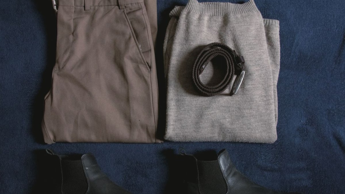 Men's clothing ensemble including trousers, sweater, boots, and belt arranged on a blue surface.