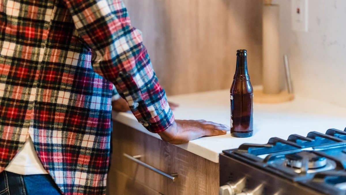 Person in plaid shirt leaning on kitchen counter with beer bottle visible.