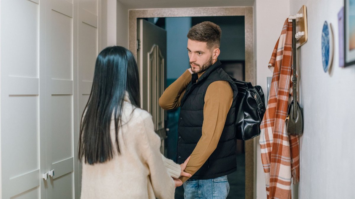 A couple holds hands in a hallway as a man prepares to leave with a backpack.