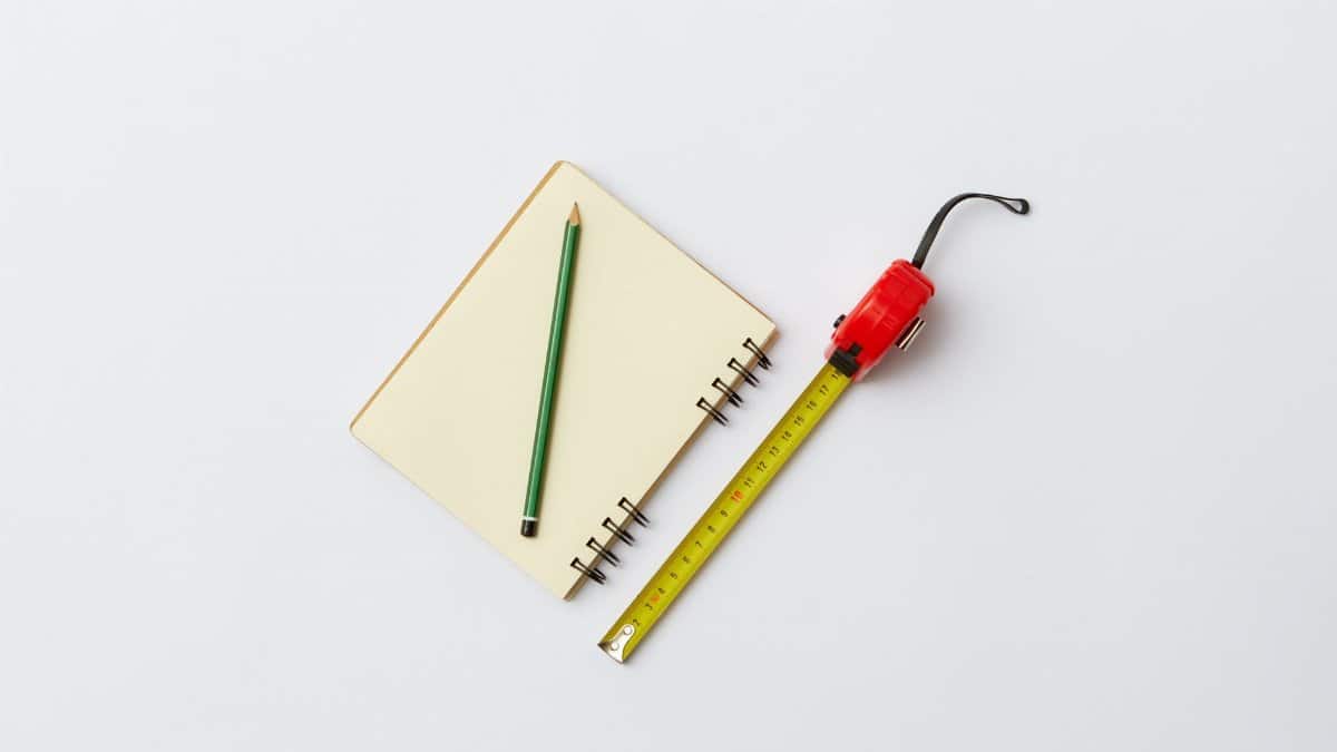 Simple flat lay of a notebook, green pencil, and red tape measure on a white background.