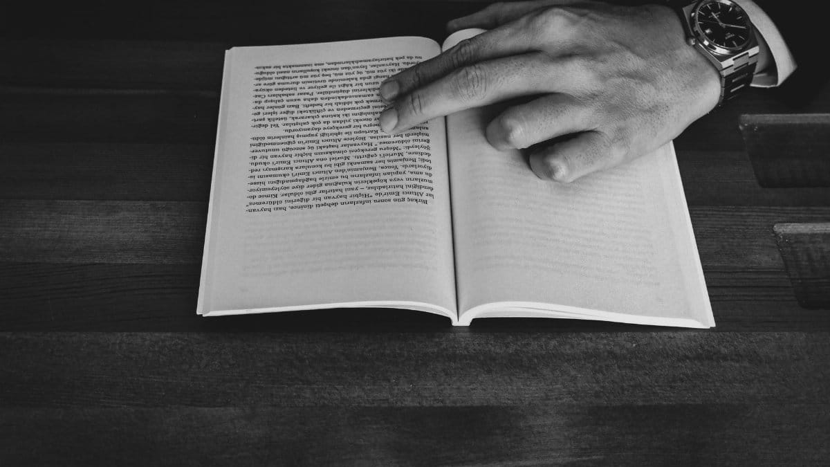 A monochrome photograph of a man's hand pointing at a book on a wooden table.