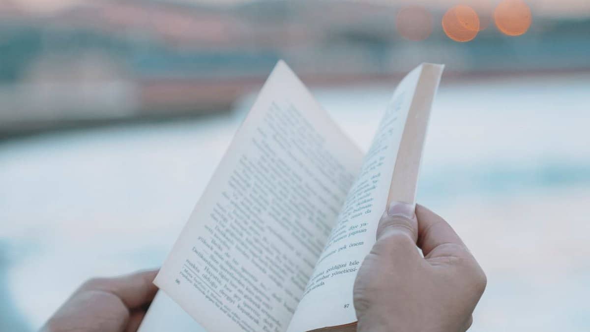 A person reading a book by the water during twilight, with blurred background lights.