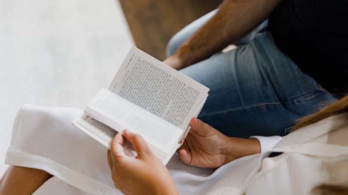 Two people reading a book together, one wearing a white dress, indoors.