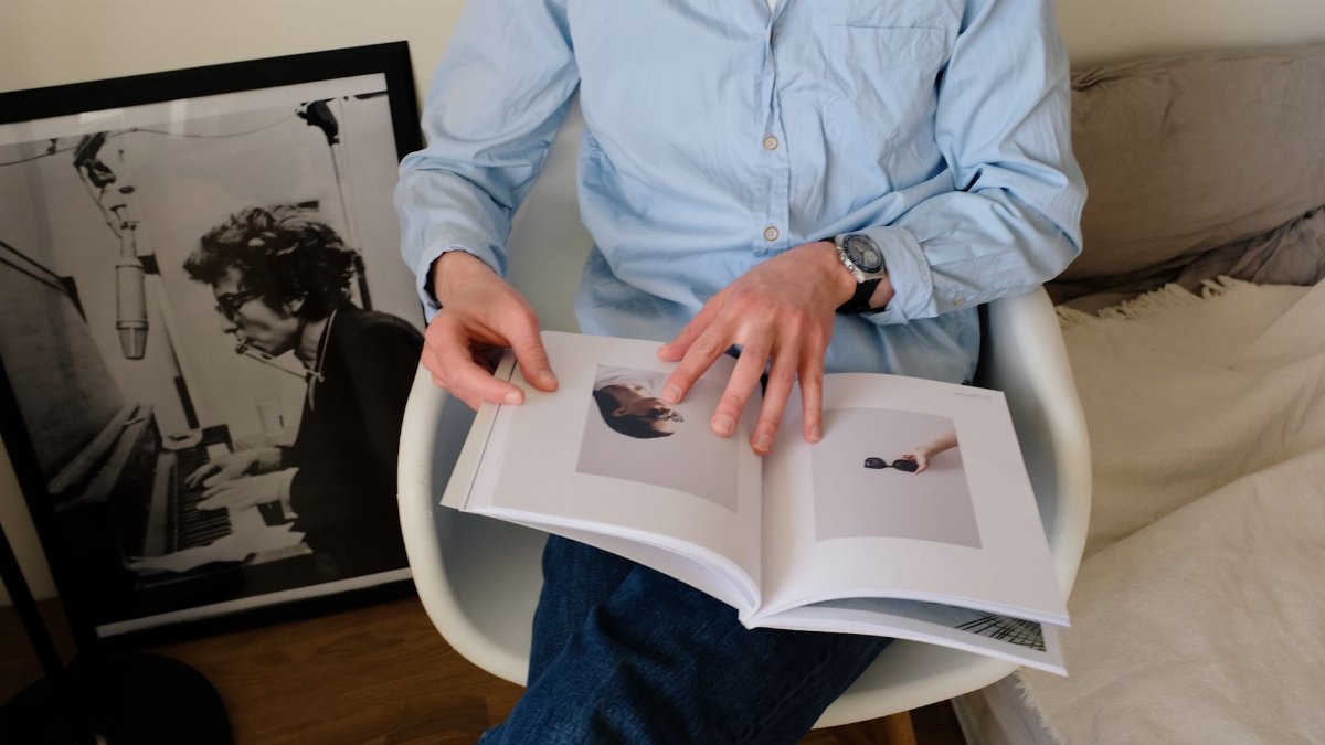 From above of crop male in casual wear sitting on chair near bed and looking through opened magazine with pictures