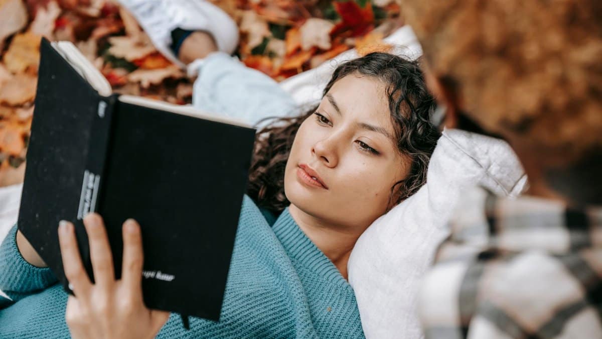 From above of pensive Hispanic female reading book while lying on lap of crop person while spending free time together in autumn nature