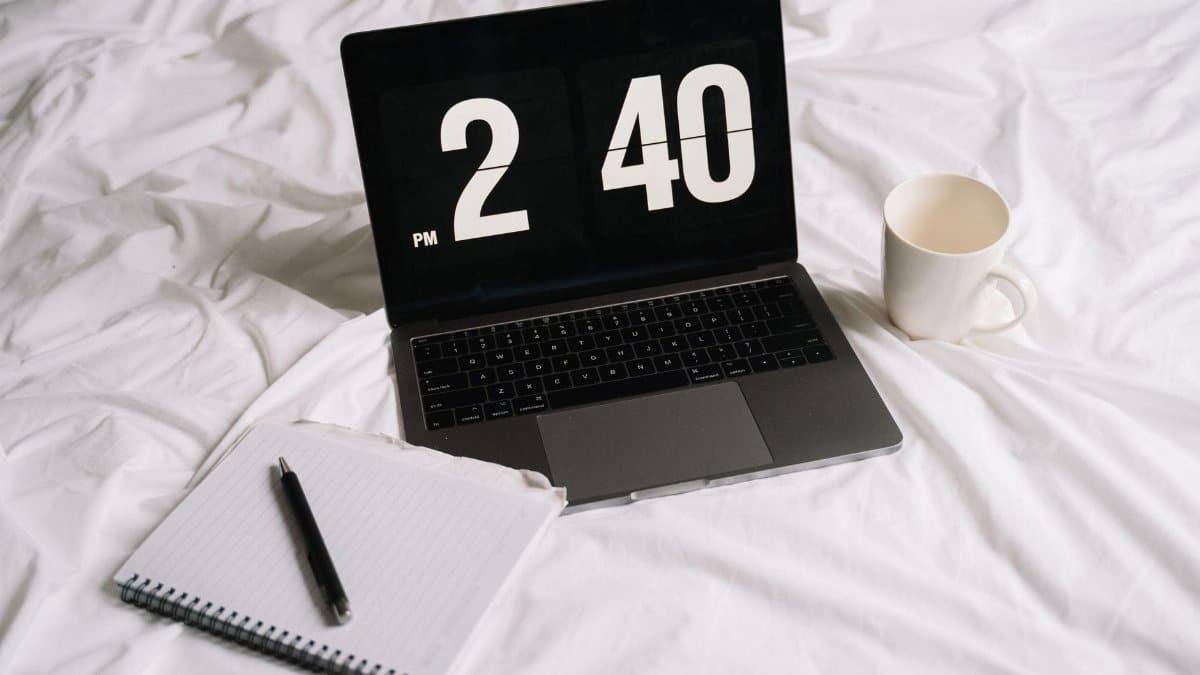 Laptop displaying time next to a notebook and coffee on a bed, creating a cozy work setup.