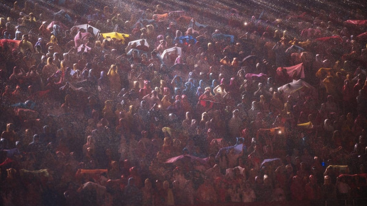 A large crowd gathers at night in the rain, illuminated by bright lights.