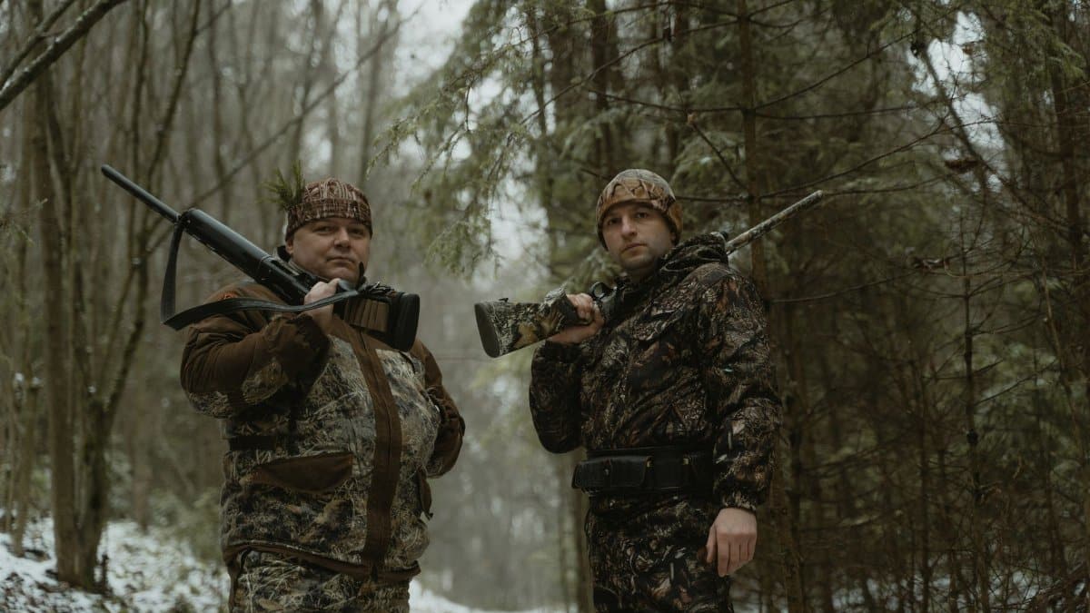 Two hunters in camouflage with rifles in a snowy forest landscape.