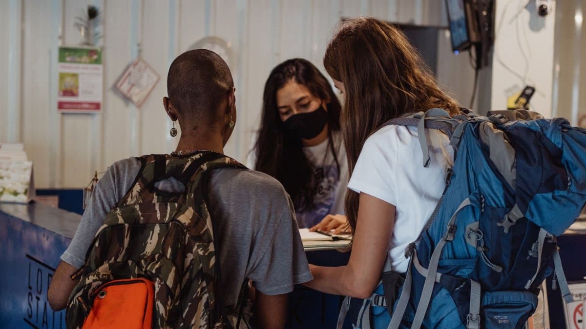 Two travelers with backpacks check in at a hostel reception counter.