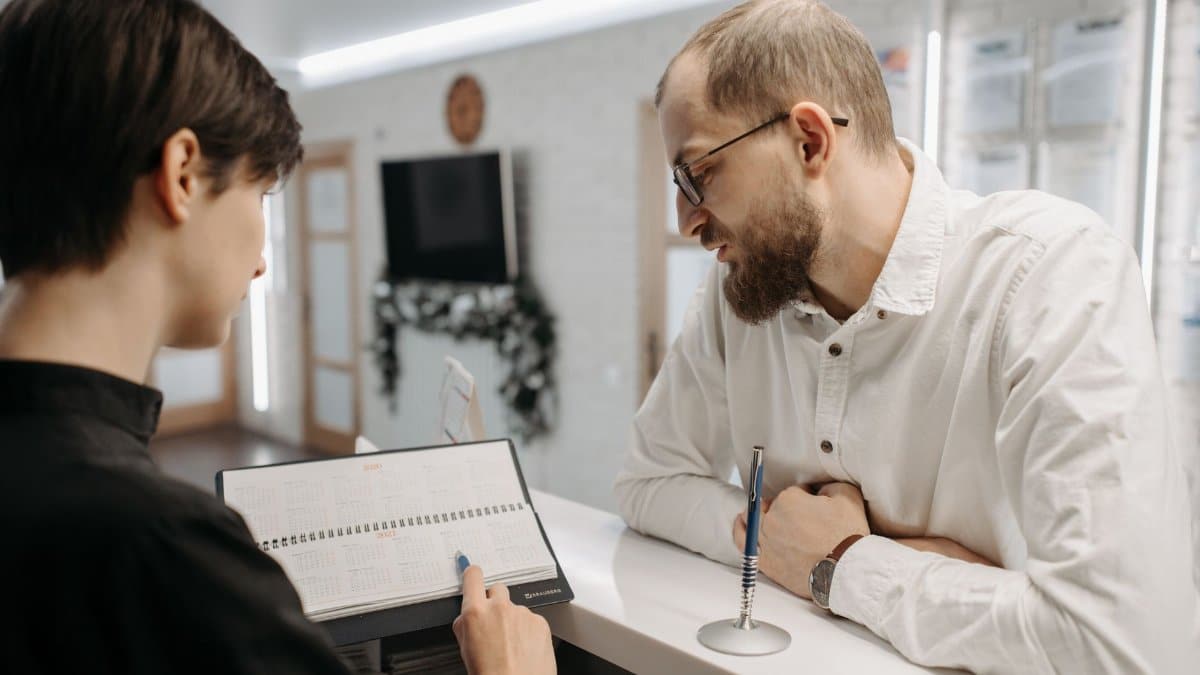 A receptionist and client converse over an appointment book at a clinic reception desk.